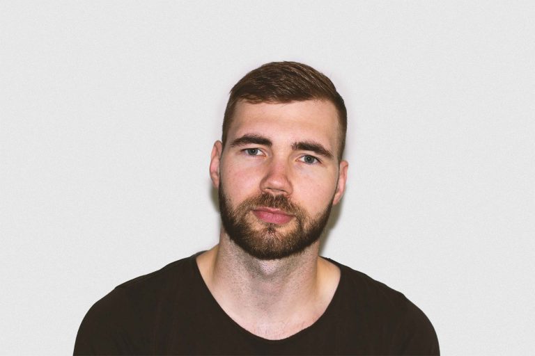 Portrait von Dominik Werner, Mann mit dunklem T-Shirt und Bart vor grauer Wand